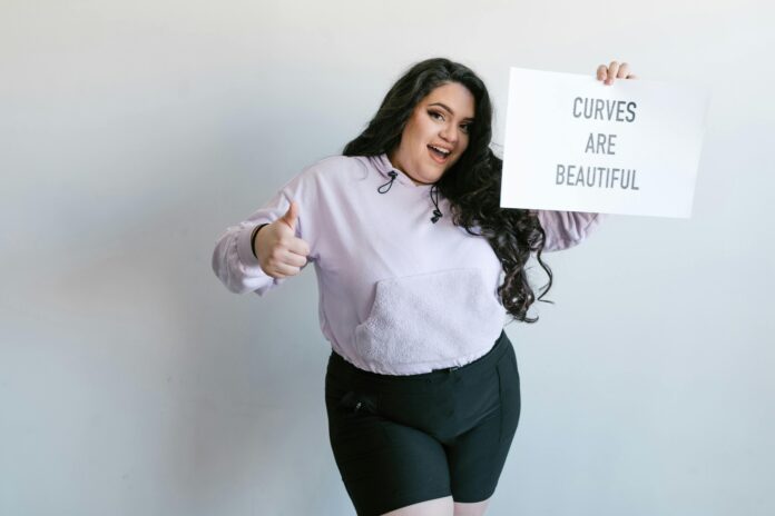 Woman wearing plus size sweatshirt and shorts holding a sign that says curves are beautiful.