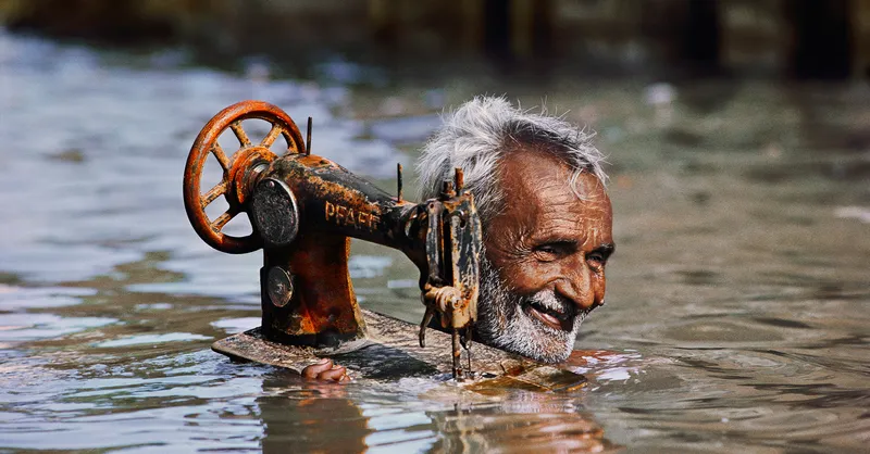 Old Indian man carrying a rusty singer sewing machine on his shoulder through floodwaters which are up to his neck.